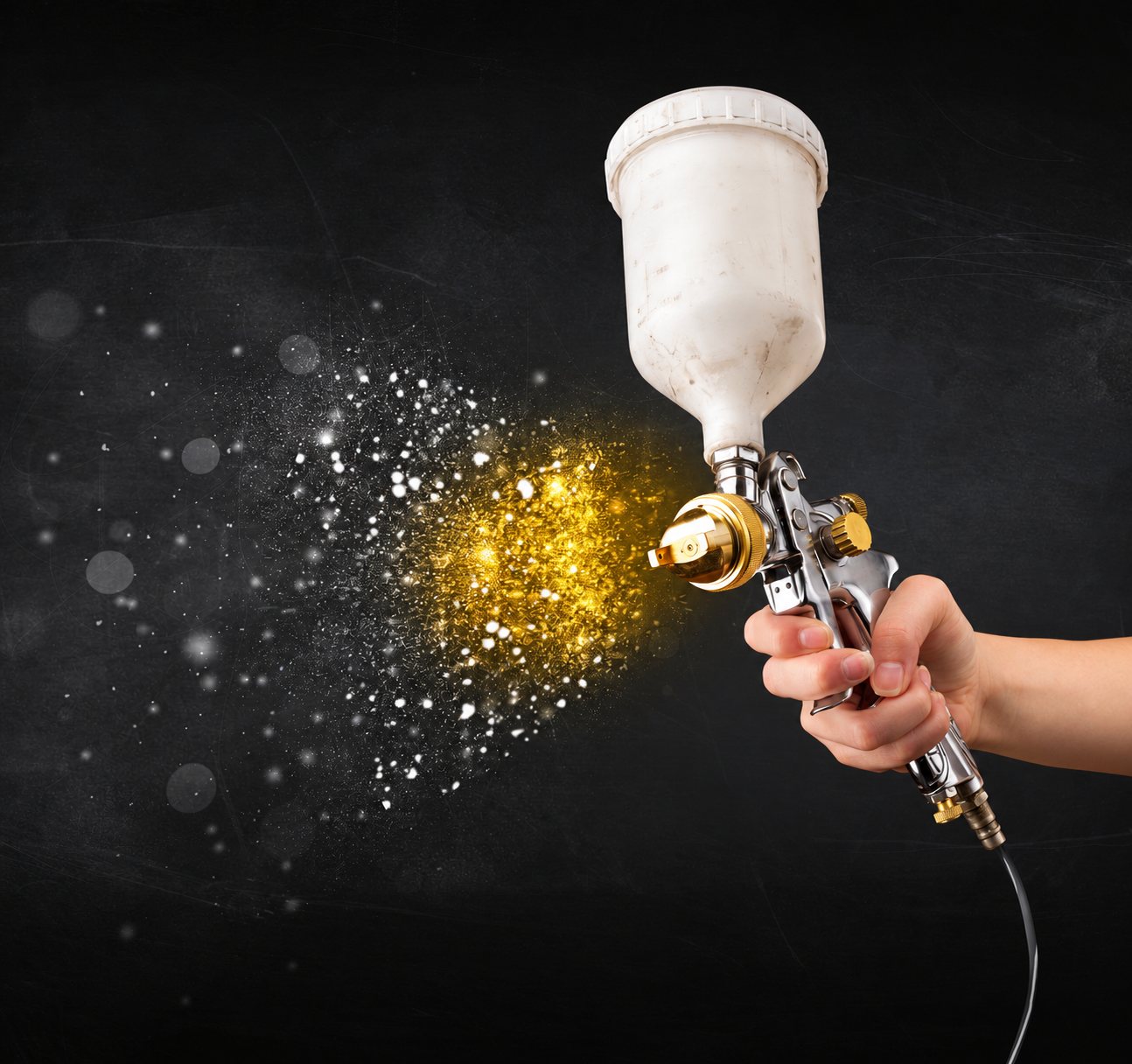Une main tient un pistolet à peinture au godet blanc qui projette un nuage de particules dorées et brillantes sur un fond sombre.